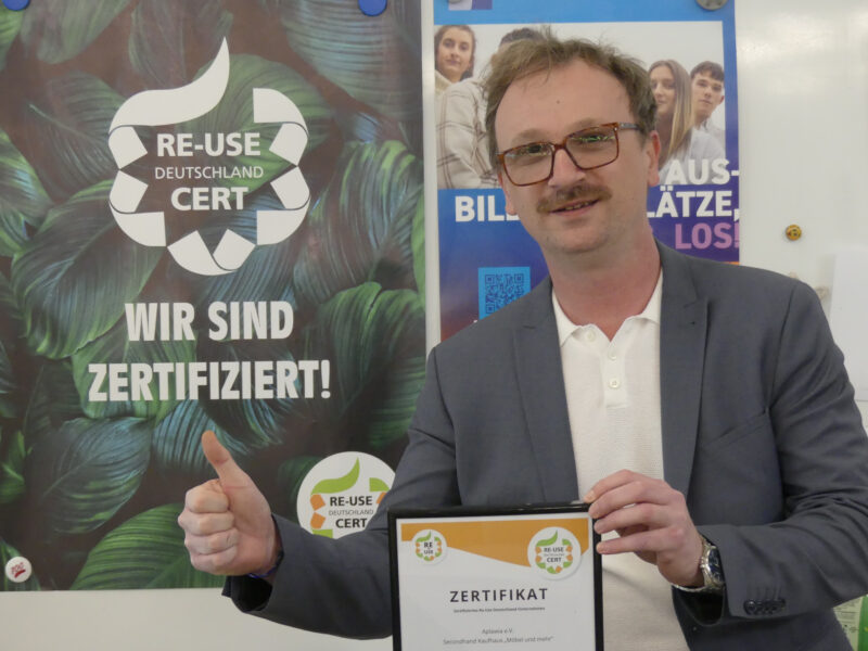 Sebastian Därr lächelt mit Daumen-hoch-Geste in die Kamera und hält das neue REUse Zertifikat. Im Hintergrund ein Plakat das 